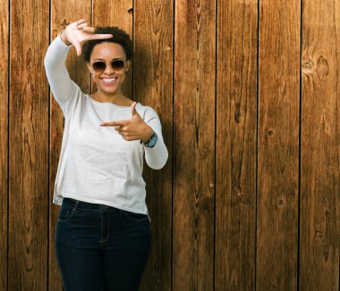 Güzel genç Afro-Amerikan kadın elle yapma çerçeve ve parmakları ile mutlu yüz gülümseyen izole arka plan üzerinde güneş gözlüğü takıyor. Yaratıcılık ve Fotoğrafçılık kavramı.