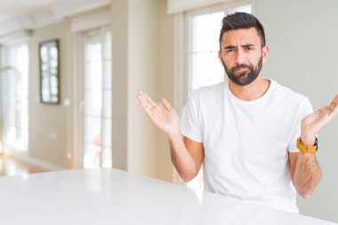 Yakışıklı İspanyol adam evde rahat beyaz t-shirt silah ve elleri kaldırdı ile clueless ve karışık ifade. Şüphe kavramı.