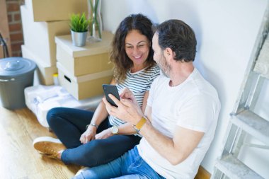 Orta yaş üst düzey romantik çift aşk etrafında kutuları ile daire katında oturan ve akıllı telefon yeni bir eve taşımak için mutlu gülümseyen kullanarak