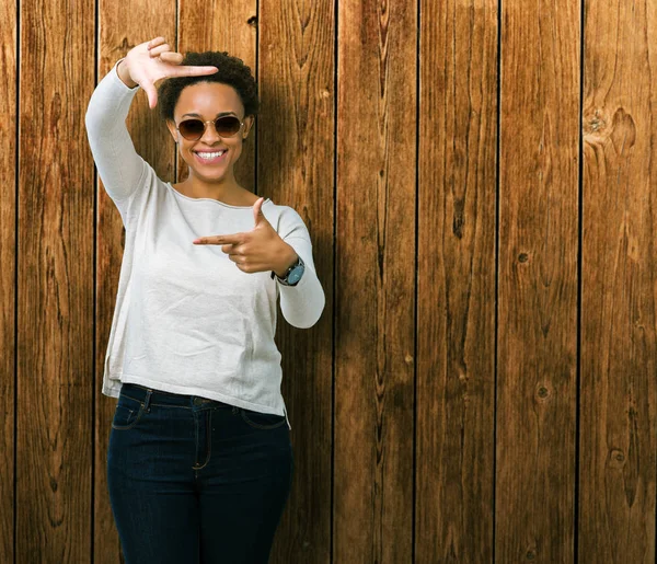 Güzel genç Afro-Amerikan kadın elle yapma çerçeve ve parmakları ile mutlu yüz gülümseyen izole arka plan üzerinde güneş gözlüğü takıyor. Yaratıcılık ve Fotoğrafçılık kavramı.
