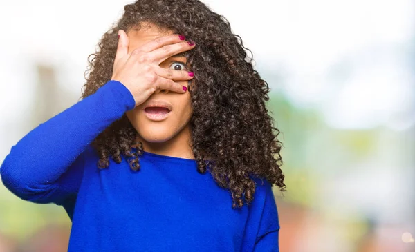 Kıvırcık saçlı kış kazak giyen genç güzel kadın şok yüz ve gözleri el ile kaplayan gözetleme, utanmış ifade ile parmaklarından bakarak.