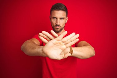 Genç yakışıklı adam kırmızı izole arka plan reddi ifade geçiş kolları ve avuç içi üzerinde rahat t-shirt giyen olumsuz işaret yapıyor, kızgın yüz