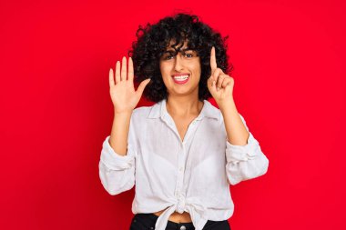 İzole kırmızı arka plan üzerinde beyaz rahat gömlek giyen kıvırcık saçlı genç arap kadın gösteren ve emin ve mutlu gülümserken parmakları ile işaret altı.