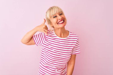 Orta yaş kadın çizgili t-shirt izole pembe arka plan üzerinde duran giyen telefonda konuşurken gibi el ve parmakları ile telefon jest yapıyor gülümseyerek. İletişim kavramları.