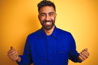 Izole sarı arka plan üzerinde üniforma giyen yakışıklı Hintli işçi adam şaşırmış ve kollarını kaldırdı ve açık gözleri ile başarı için şaşırttı. Kazanan kavramı.