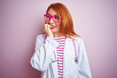 Genç redhead doktor kadın izole pembe arka plan üzerinde duran stetoskop kullanarak stresli ve ağız tırnak ısırma elleri ile sinir görünümlü. Anksiyete sorunu.