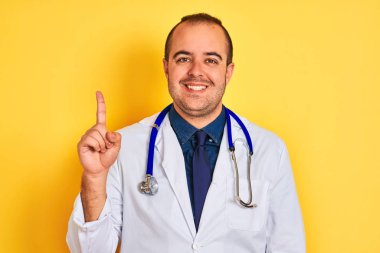 Genç doktor adam ceket ve stetoskop izole sarı arka plan üzerinde duran gösteren ve kendinden emin ve mutlu gülümserken parmak sayısı bir işaret giyen.