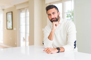 Evde rahat beyaz kazak giyen İspanyol yakışıklı emin çapraz kollar ve çeneme kaldırdı el ile gülümseme ile kamera arıyorum. Pozitif düşünce.