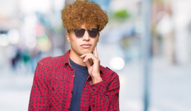 Yakışıklı genç afro saçlı çene düşünme hakkında soru, dalgın ifade üzerine el ile güneş gözlüğü takıyor. Düşünceli yüzüyle gülümsüyor. Şüphe kavramı.