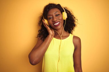 Afro-Amerikan kadın mutlu bir yüz ayakta ve dişleri gösteren kendinden emin bir gülümseme ile gülümseyerek izole sarı arka plan üzerinde kulaklık kullanarak müzik dinlerken