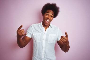 Afro saçlı beyaz gömlek giyen genç amerikalı adam izole pembe arka plan üzerinde duran deli ifade elleri ile rock sembolü yapıyor bağırarak. Müzik yıldızı. Ağır konsept.