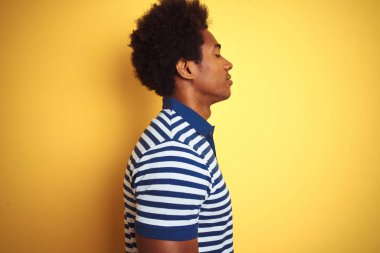 Afro saçlı afro saçlı amerikan adam yan bakarak izole sarı arka plan üzerinde duran lacivert çizgili polo giyen, kendine güvenen gülümseme ile doğal yüz ile profil poz dinlenmek.