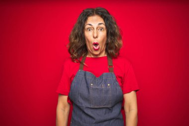 Orta yaş yaşlı kadın kırmızı izole arka plan üzerinde önlük üniforma giyen korkmuş ve sürpriz ve şaşkın ifade, korku ve heyecanlı yüz ile şok.