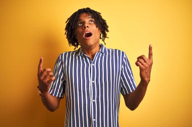 Izole sarı arka plan üzerinde rahat çizgili t-shirt giyen dreadlocks ile Afro adam şaşırttı ve yukarı bakarak ve parmakları ile işaret ve kolları kaldırdı şaşırttı.