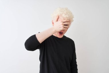 Genç albino sarışın adam siyah t-shirt izole beyaz arka plan üzerinde duran şok el ile yüz ve gözleri kapsayan gözetleme, utanmış ifade ile parmaklarından bakarak giyiyor.