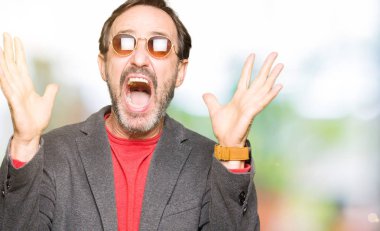 Orta yaş yakışıklı iş adamı deli kutluyor ve silah ile başarı için heyecanlı çığlık kaldırdı ve açık gözleri hayret güneş gözlüğü takıyor. Kazanan kavramı