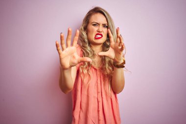 Pembe izole arka plan üzerinde duran t-shirt giyen genç güzel kadın korkmuş ve korku ifade ile korkmuş elleriyle jest durdurmak, şok içinde bağırarak. Panik kavramı.