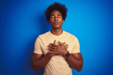 Afro saçlı Amerikalı adam izole mavi arka plan üzerinde çizgili sarı t-shirt giyen kapalı gözleri ve yüzünde minnettar jest göğüs elleri ile gülümseyerek. Sağlık kavramı.