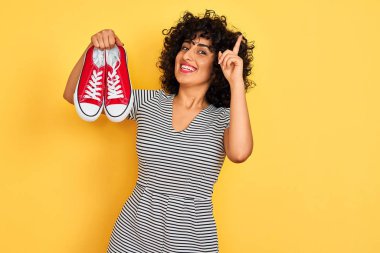 İzole sarı arka plan üzerinde rahat spor ayakkabı tutan kıvırcık saçlı genç arap kadın mutlu yüz, bir numara ile bir fikir veya soru işaret parmağı ile şaşırttı