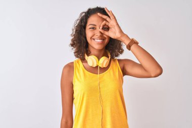İzole beyaz arka plan üzerinde sarı kulaklık takan genç Brezilyalı kadın el gülümseyerek ok jest yapıyor, mutlu yüz ile parmaklardan bakan göz.