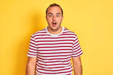 Izole sarı arka plan üzerinde duran rahat çizgili t-shirt giyen genç adam korkmuş ve sürpriz ifade, korku ve heyecanlı yüz ile şok.