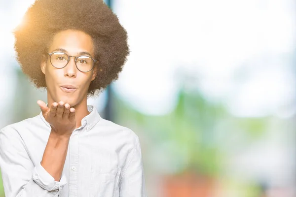 Young african american man with afro hair wearing glasses looking at the camera blowing a kiss with hand on air being lovely and sexy. Love expression.