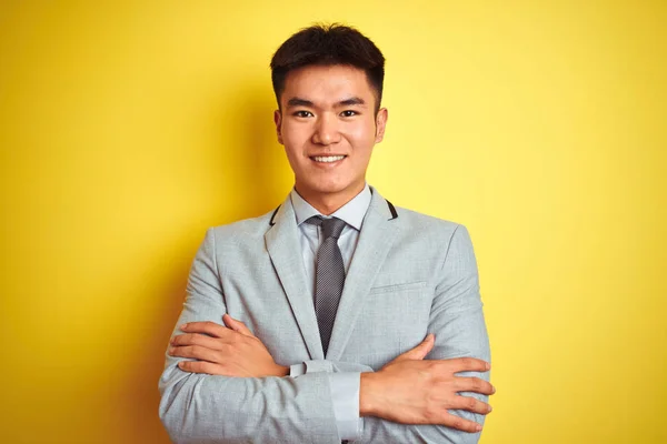 Asian chinese businessman wearing suit and tie standing over isolated yellow background happy face smiling with crossed arms looking at the camera. Positive person.