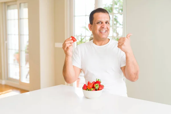 Middle age man eating strawberries at home pointing and showing with thumb up to the side with happy face smiling