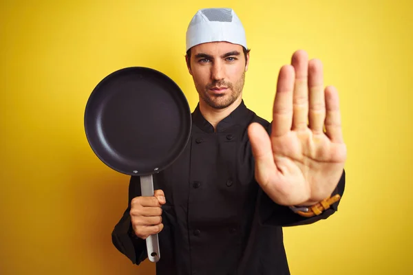 年轻的厨师男子穿着制服和帽子拿着厨师锅在孤立的黄色背景与打开的手做停止标志与认真和自信的表情 防御手势 — 图库照片