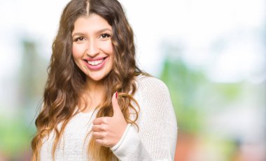 Beyaz kazak giyen genç güzel kadın eliyle jest kadar mutlu başparmak yapıyor. Başarı gösteren kameraya bakarak ifadeyi onaylama.