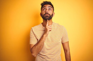 İzole sarı arka plan üzerinde duran t-shirt giyen genç hintli adam çene üzerinde parmak ile şüphe hakkında konsantre ve merak bakarak