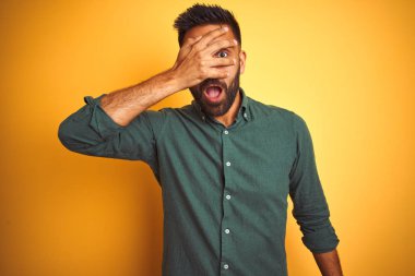 Genç hintli işadamı izole beyaz arka plan üzerinde duran zarif gömlek giyen şok yüz ve gözleri el ile kaplayan gözetleme, utanmış ifade ile parmaklarından bakarak.