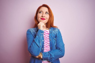 Denim gömlek giyen güzel kızıl saçlı kadın ve izole pembe arka plan üzerinde çizgili t-shirt çene üzerinde el soru, dalgın ifade düşünme ile. Gülümseyen ve düşünceli bir yüz. Şüphe kavramı.