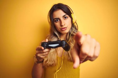 Genç güzel kadın kamera ve size parmak ile işaret sarı izole arka plan üzerinde gamepad kullanarak video oyunu oynarken, el işareti, önden olumlu ve kendinden emin jest