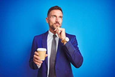 Genç yakışıklı iş adamı mavi izole arka plan ciddi yüz soru hakkında düşünme, çok karışık bir fikir üzerinde bir kağıt fincan üzerinde bir kahve içme