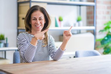 Orta yaş kıdemli kadın masada oturan evde gülümseyen ve iki el ve parmak tarafına işaret kamera bakıyor.