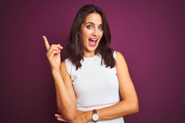 Yüzünde büyük bir gülümseme ile mor izole arka plan üzerinde duran beyaz elbise giyen genç güzel kadın, kameraya bakarak yan el ve parmak ile işaret.