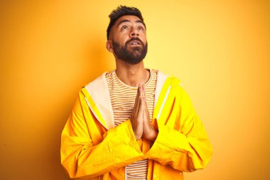 Yağmurluk giyen genç hintli adam izole sarı arka plan üzerinde ayakta dilenmek ve elleri ile birlikte dua umut ifadesi ile yüzünde çok duygusal ve endişeli. Af dilemek istiyorum. Din kavramı.