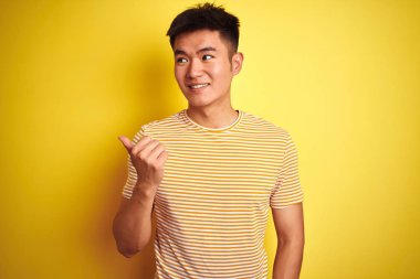 İzole sarı arka plan üzerinde duran t-shirt giyen genç Asyalı Çinli adam mutlu yüz görünümlü gülümseyerek ve başparmak ile yan işaret.