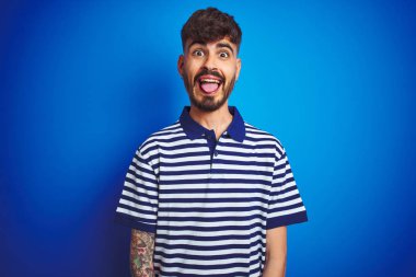 Çizgili polo giyen dövme giyen genç adam komik ifade ile mutlu dil yapışmasını izole mavi arka plan üzerinde duran. Duygu kavramı.