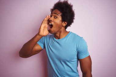 Afro saçlı afro saçlı, izole edilmiş pembe arka planın üzerinde duran mavi tişört giyen Afro-Amerikan adam bağırarak ve ağızda el ile yan yüksek sesle bağırıyor. İletişim kavramı.