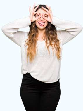 Beyaz kazak giyen genç güzel kadın dürbün gibi tamam jest yapıyor dilini dışarı sokuyor, gözleri parmaklardan bakıyor. Çılgın ifade.