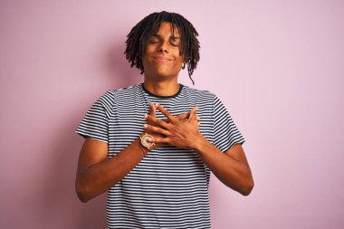 Dreadlocks lacivert çizgili t-shirt kapalı gözleri ve yüzünde minnettar jest ile göğüs üzerinde elleri ile gülümseyerek izole pembe arka plan üzerinde duran giyen Afro adam. Sağlık kavramı.