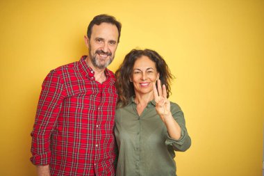 Izole sarı arka plan üzerinde Güzel orta yaş çift gösteren ve emin ve mutlu gülümserken parmakları sayısı üç işaret.