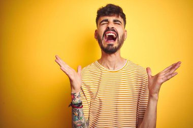 Dövme ile genç adam izole sarı arka plan üzerinde duran çizgili t-shirt dikilmiş silah ile başarı için deli ve çılgın kutluyor ve kapalı gözleri heyecanlı çığlık kaldırdı. Kazanan kavramı