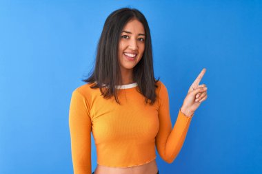 Turuncu tişört giyen genç güzel Çinli kadın yüzünde büyük bir gülümseme ile izole mavi arka plan üzerinde duran, kameraya bakarak yan el ve parmak ile işaret.