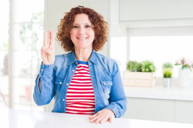 Kıvırcık saçlı orta yaş yaşlı kadın evde kot ceket giyen gösteren ve parmakları ile işaret ederken emin ve mutlu gülümseyerek üç numara.