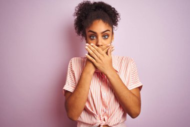 Izole pembe arka plan üzerinde duran rahat çizgili gömlek giyen Afro-Amerikan kadın hata için elleri ile ağız kapsayan şok. Gizli kavram.
