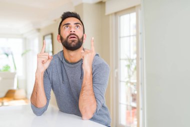 Yakışıklı İspanyol adam evde rahat sweatshirt giyen şaşırttı ve yukarı bakarak şaşırttı ve parmakları ile işaret ve kolları kaldırdı.