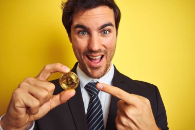 İzole sarı arka plan üzerinde duran bitcoin tutan genç yakışıklı işadamı el ve parmak ile işaret çok mutlu